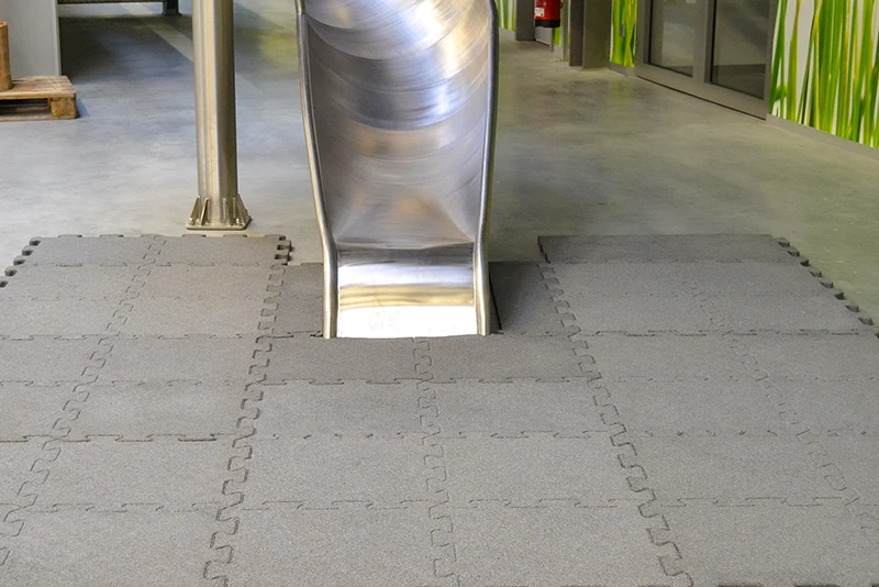 Le bas d’un toboggan en acier inoxydable installé en intérieur, avec un sol recouvert de dalles en caoutchouc antichoc grises de WARCO. Ces dalles sont emboîtées grâce à un système puzzle, assurant une fixation stable et sécurisée. Leur surface amortissante réduit les risques de blessure en cas de chute, ce qui les rend idéales pour les aires de jeux intérieures et les salles d’escalade. En arrière-plan, un décor végétal apporte une ambiance naturelle à l’espace.