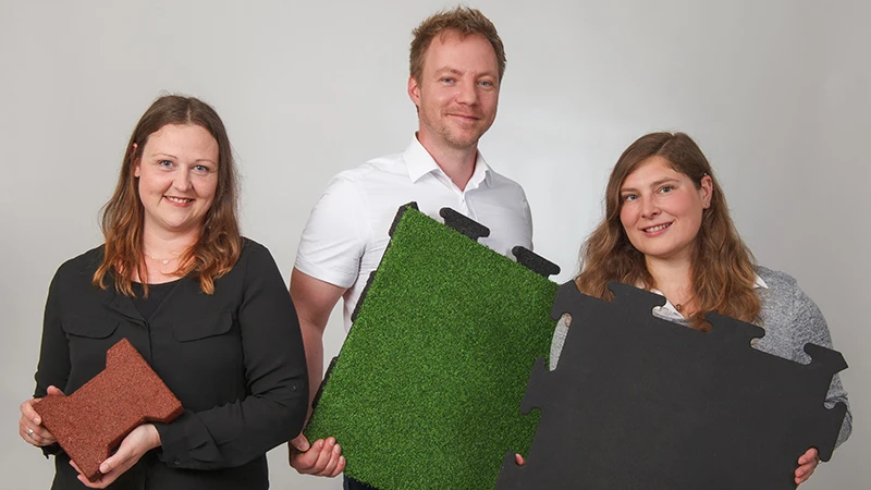 Souriants, trois conseillers WARCO présentent des dalles en granulat de caoutchouc lié au PU, déclinées en brun, vert et noir. Conçues pour le balcon, elles offrent une surface antidérapante et résistante. Chaque modèle possède un design unique et s’installe facilement. Grâce à leur expertise, vous trouverez le revêtement idéal pour un balcon sûr et esthétique. Faciles d’entretien, ces dalles apportent également un grand confort.