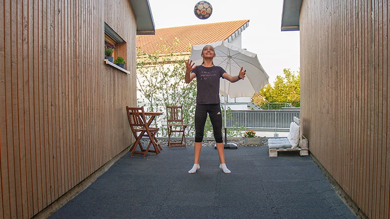 Une fille joue au ballon sur une terrasse entre deux murs en bois. Le sol est composé de dalles de terrasse en granulat de caoutchouc noir, offrant une surface robuste et élastique, adaptée aux activités sportives. En arrière-plan, un parasol et un petit ensemble de sièges en bois créent une ambiance accueillante.