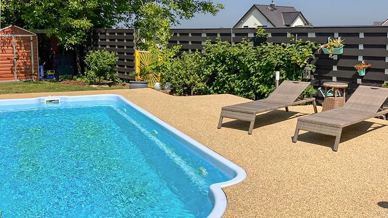 Privater Pool mit beiger Umrandung, Liegestühlen und üppiger Bepflanzung: Die WARCO Poolumrandung ist rutschhemmend, angenehm barfuß begehbar, hitzebeständig und pflegeleicht – ideal für sicheres Baden und entspanntes Sonnen.