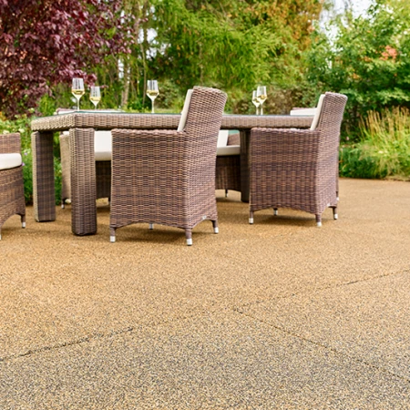 Terrasse baignée de soleil dans un jardin, équipée de dalles WARCO beige-brun en granulats de caoutchouc liés au PU. La surface antidérapante et perméable à l’eau assure un confort de marche optimal et atténue les bruits. Une table de jardin robuste avec chaises tressées et verres de vin remplis invite à la détente. Arbustes luxuriants et fleurs colorées créent une atmosphère naturelle. Ce revêtement élastique résiste au gel, conserve sa forme et convient à un usage toute l’année.