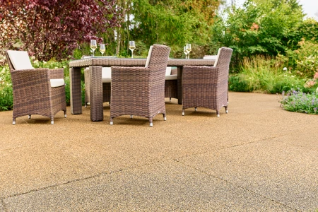Terrasse baignée de soleil dans un jardin, équipée de dalles WARCO beige-brun en granulats de caoutchouc liés au PU. La surface antidérapante et perméable à l’eau assure un confort de marche optimal et atténue les bruits. Une table de jardin robuste avec chaises tressées et verres de vin remplis invite à la détente. Arbustes luxuriants et fleurs colorées créent une atmosphère naturelle. Ce revêtement élastique résiste au gel, conserve sa forme et convient à un usage toute l’année.
