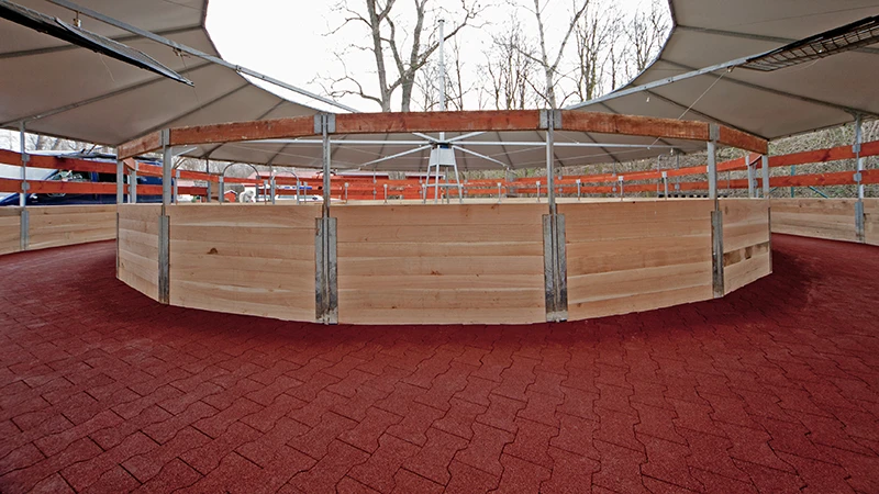 La photo montre un marcheur couvert moderne pour chevaux. La piste est entièrement pavée de dalles en caoutchouc rouge – antidérapantes, perméables et douces pour les articulations. La surface élastique permet aux chevaux de se déplacer en toute sécurité, par tous les temps et à toutes les allures. La piste est stable, durable et nécessite très peu d’entretien. L’installation convient parfaitement aux centres équestres, élevages et cliniques équines recherchant un sol adapté aux chevaux, peu contraignant et durable.