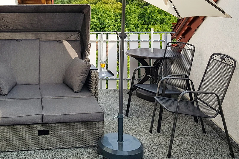 La photo montre un balcon recouvert de dalles WARCO grises. Un confortable fauteuil de plage et une petite table avec deux chaises en métal offrent un coin détente parfait. Le revêtement élastique et antidérapant protège de l’humidité et atténue le bruit des pas. Il est également facile à entretenir et résistant aux intempéries, assurant un espace extérieur accueillant sur la durée.