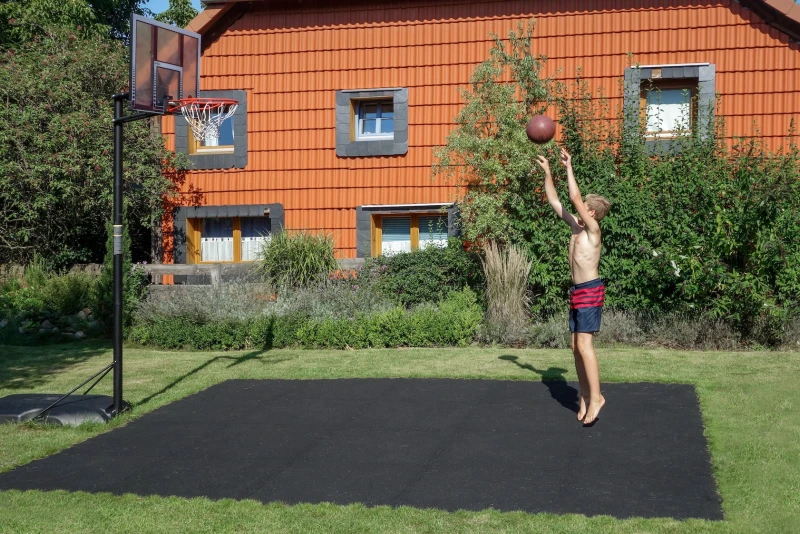 Le petit terrain de basket fait de dalles de jeu de ballon noires de WARCO a été créé par la famille elle-même dans le jardin. Un garçon joue au basket.