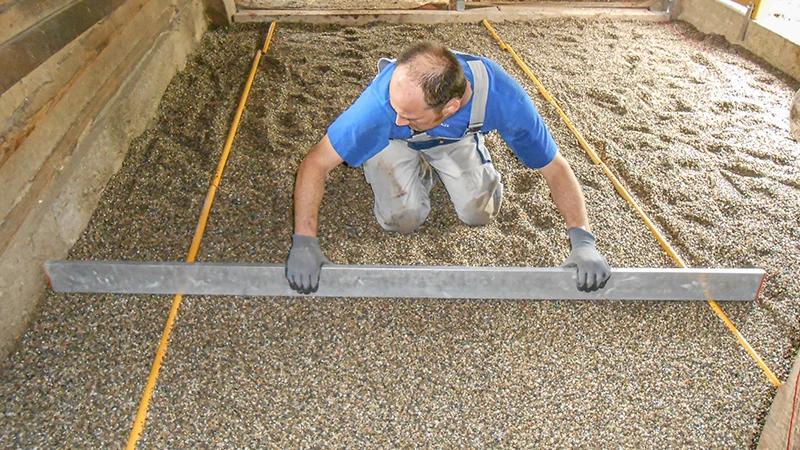 Installation d’un nouveau sol dans une écurie : Un homme utilise une règle pour niveler une couche de gravier. Le gravier sert de lit de pose pour un sol de box en granulé de caoutchouc. La zone est délimitée latéralement par des parois en bois ou en béton. Le travail est réalisé avec soin et précision – typique d’une préparation professionnelle d’un sol d’écurie adapté aux chevaux.