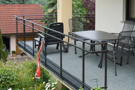 Cette photo montre un grand balcon recouvert de dalles WARCO grises. Un garde-corps en métal noir entoure la zone pour plus de sécurité. Une table spacieuse et des chaises invitent à la convivialité. Le revêtement élastique et antidérapant résiste aux intempéries et s’entretient facilement, assurant une protection durable et un espace extérieur confortable pour se détendre.
