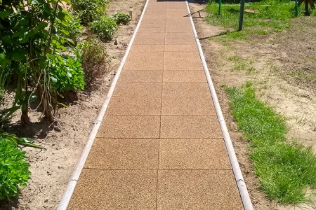 Une allée droite de jardin en dalles carrées de caoutchouc avec bordure latérale traverse le massif. Les dalles brunes en granulés de caoutchouc offrent une surface antidérapante, résistante aux intempéries et facile d’entretien. L’allée assure une marche sûre et propre par tous les temps et sépare nettement les parterres de la pelouse. Idéal pour jardins, cours et accès.