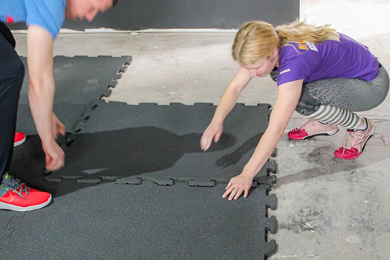 Deux personnes installent des dalles en caoutchouc antichoc grises de WARCO sur un sol en béton. Grâce au système d’emboîtement puzzle, les dalles s’assemblent facilement et sans outil. Ce revêtement amortissant est idéal pour les salles de sport, les aires de jeux et tout espace nécessitant un sol sécurisé et durable. Une solution simple et efficace pour une installation rapide et une protection optimale.